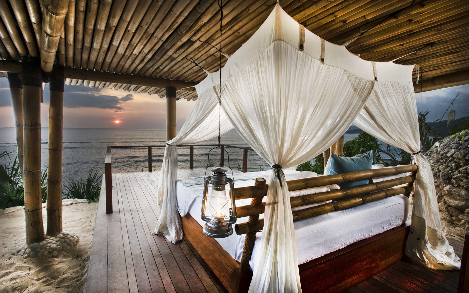 A four-poster open to the Indian Ocean at sunrise, sand underfoot, a lantern hung from the bamboo roof