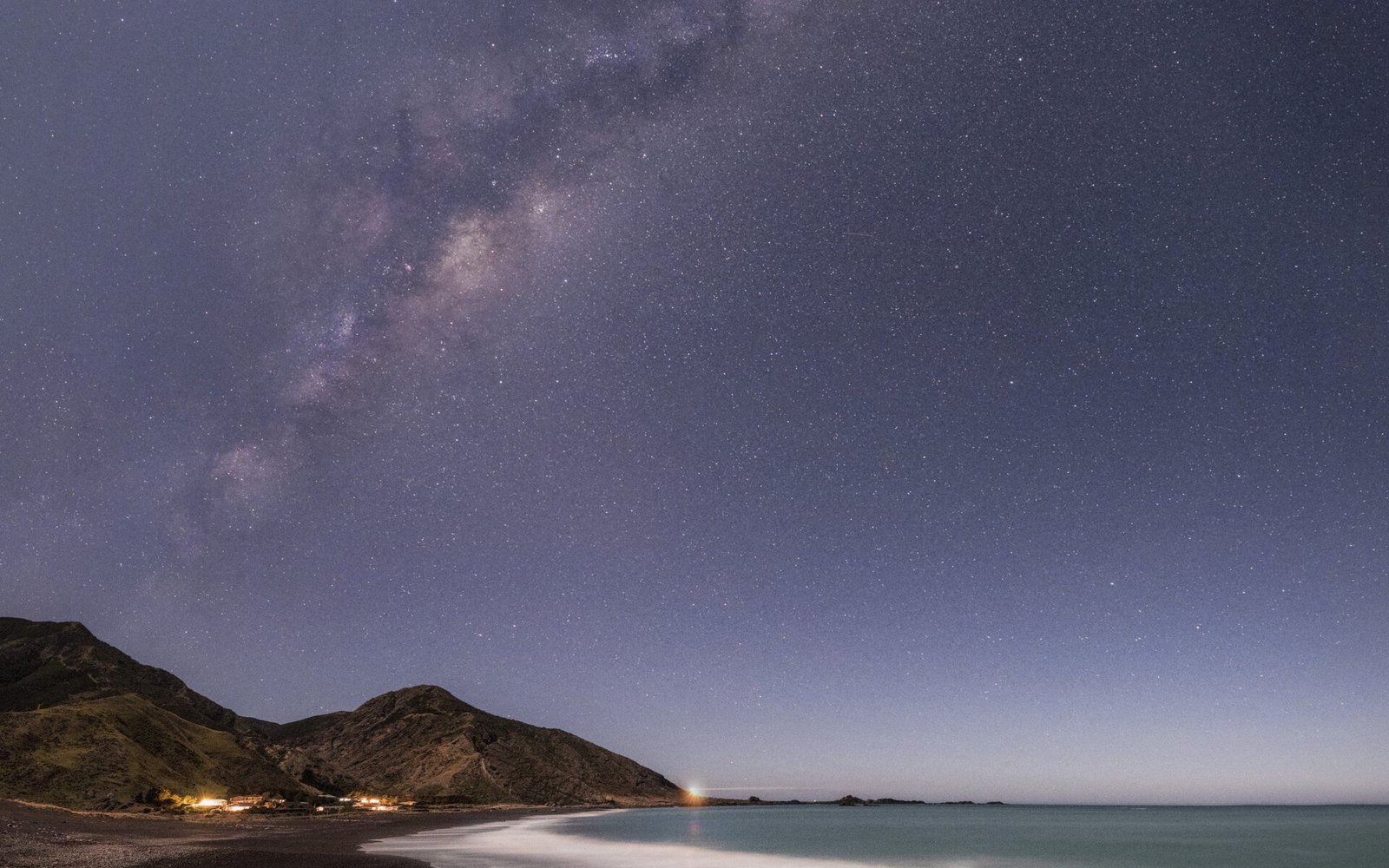 The Milky Way over Palliser Bay, a small cluster of lights at the foot of the hills, the surf drawn into a white band by the long exposure