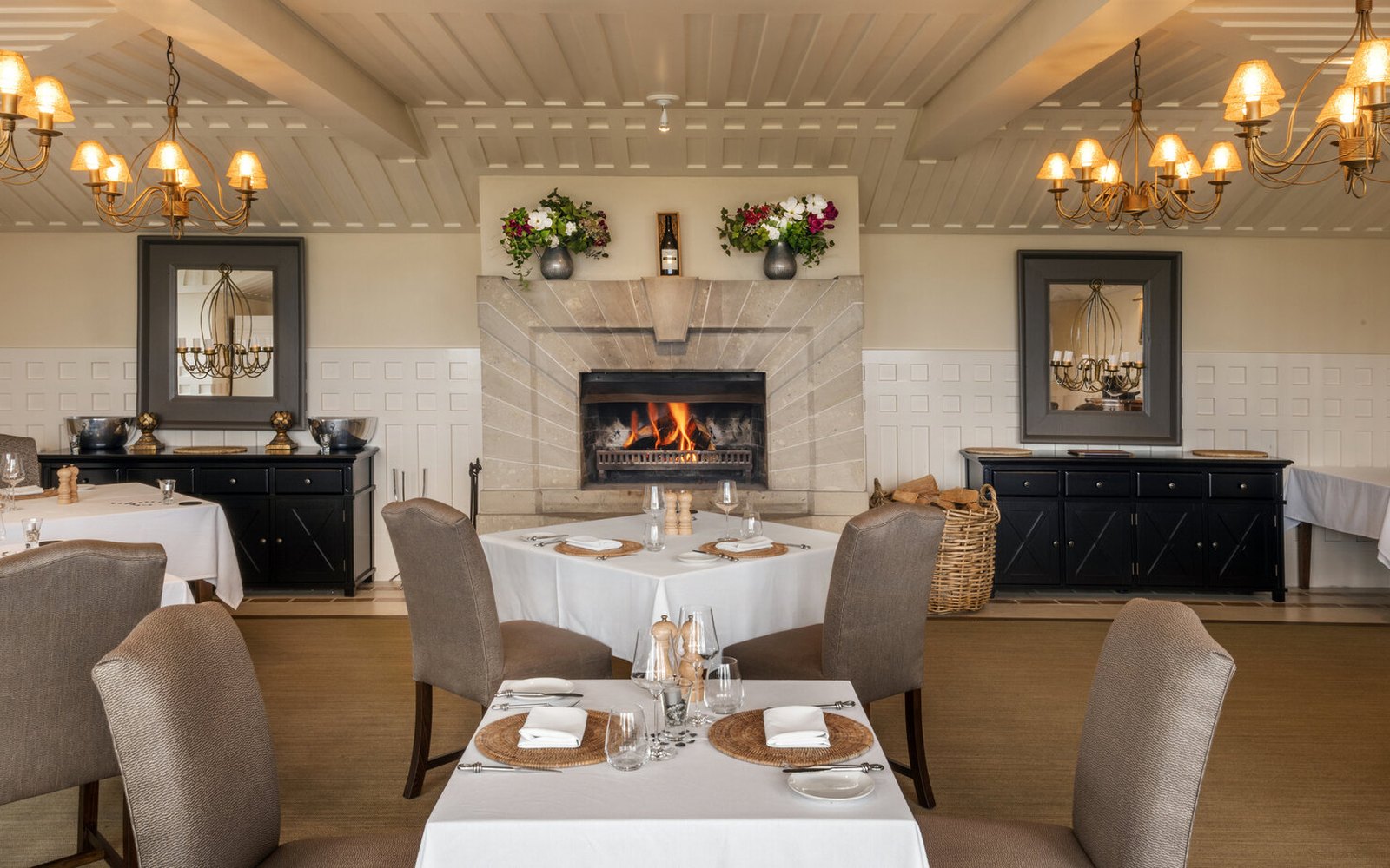 The Dining Room with its fire lit, brass chandeliers either side, white linen and woven mats on the tables