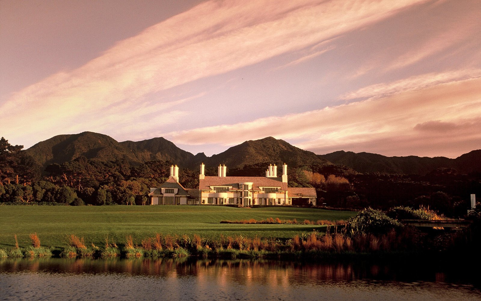 The lodge at dusk, tall chimneys above the roofline, lawn and a pond in front, the Remutaka ridge behind