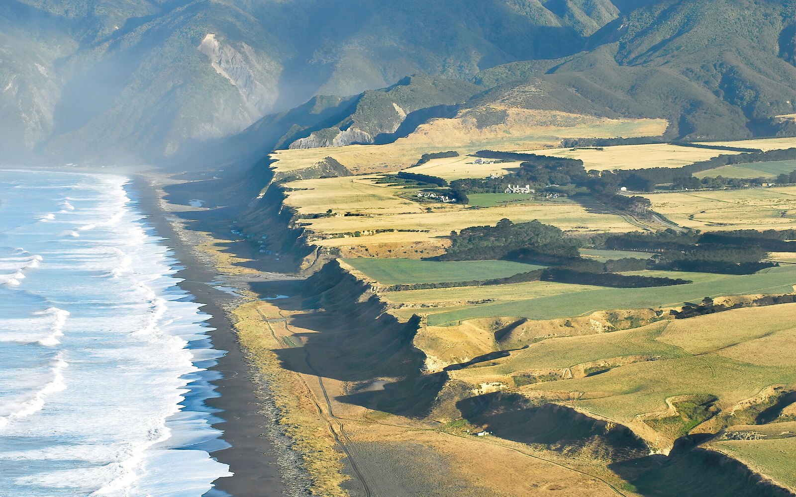 The estate seen from the air: the black sand beach and surf line of Palliser Bay on one side, farmland cut off at the cliff edge on the other, the white lodge on the terrace between them