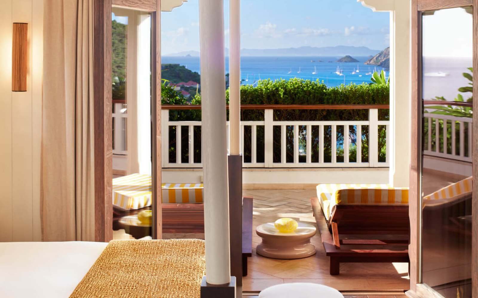 From inside a room: a four-poster corner, double doors open onto a private terrace with striped loungers, and the bay beyond