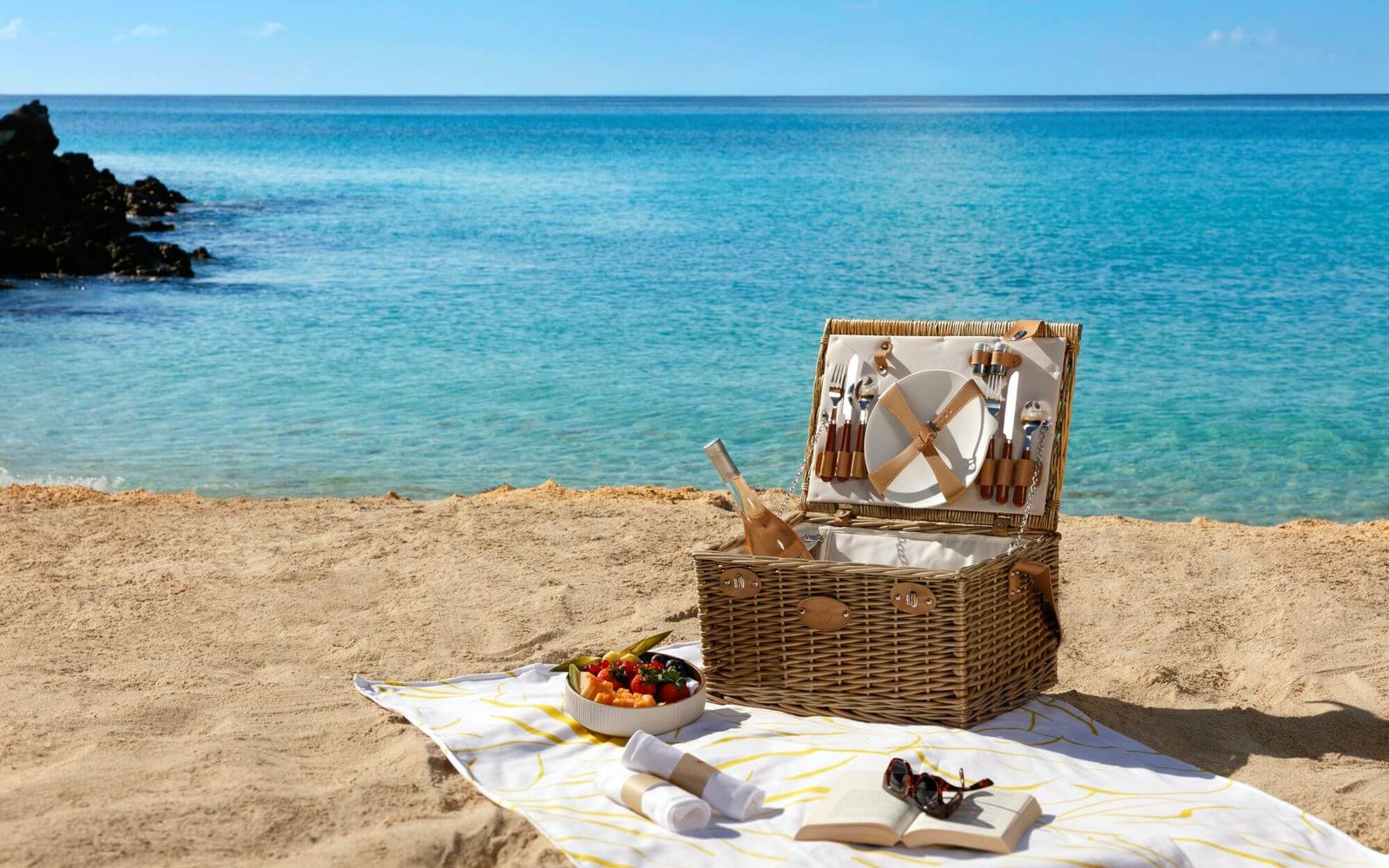 Official hotel image: an open wicker picnic hamper on a yellow-striped cloth, turquoise shallows and a black rock behind