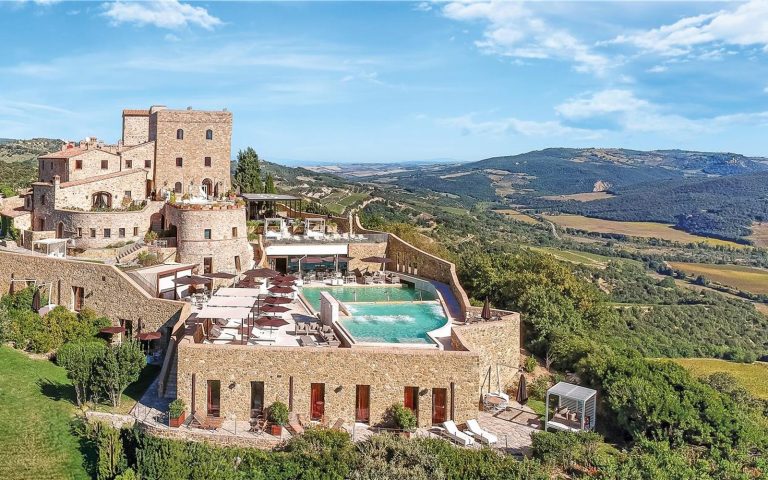 Castello di Velona 全景：石砌城堡立于蒙塔尔奇诺山脊，露天温泉池外是奥尔恰谷的丘陵与葡萄园